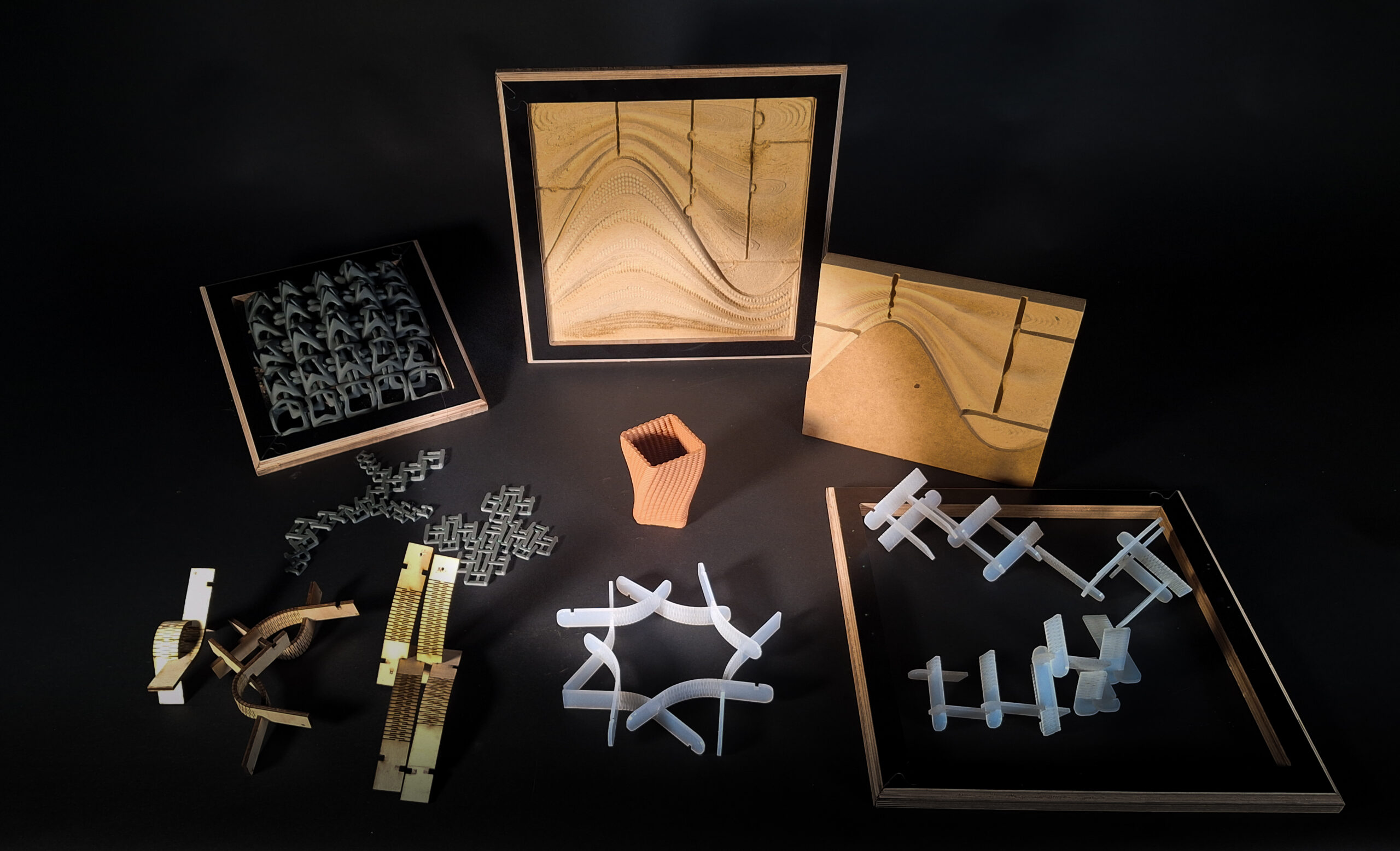 A collection of digital fabrication prototypes displayed on a dark background, including 3D-printed modular façade components, laser-cut bending assemblies, CNC-milled MDF panels with carved topographies, and a robotically 3D-printed clay vessel, all arranged within wooden frames and scattered test pieces.